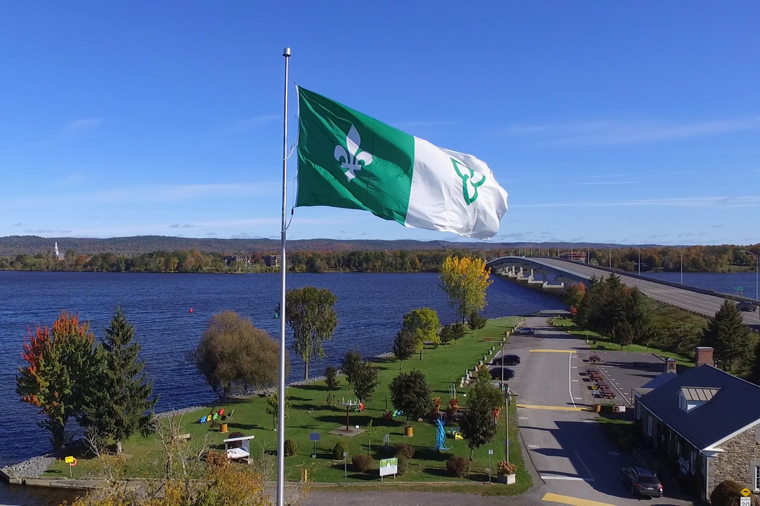 An aerial view of Confederation Park in Hawkesbury on the Ottawa River in autumn colour, with the green and white Franco-Ontarian flag in the foreground and a bridge across the river behind it.